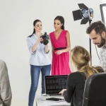 Beyaz arka planlı bir stüdyoda, bir fotoğrafçı modelle çekim detaylarını tartışıyor. Arka planda ekip üyeleri bilgisayar üzerinden görselleri inceliyor ve ışık ekipmanları çekim ortamını tamamlıyor.